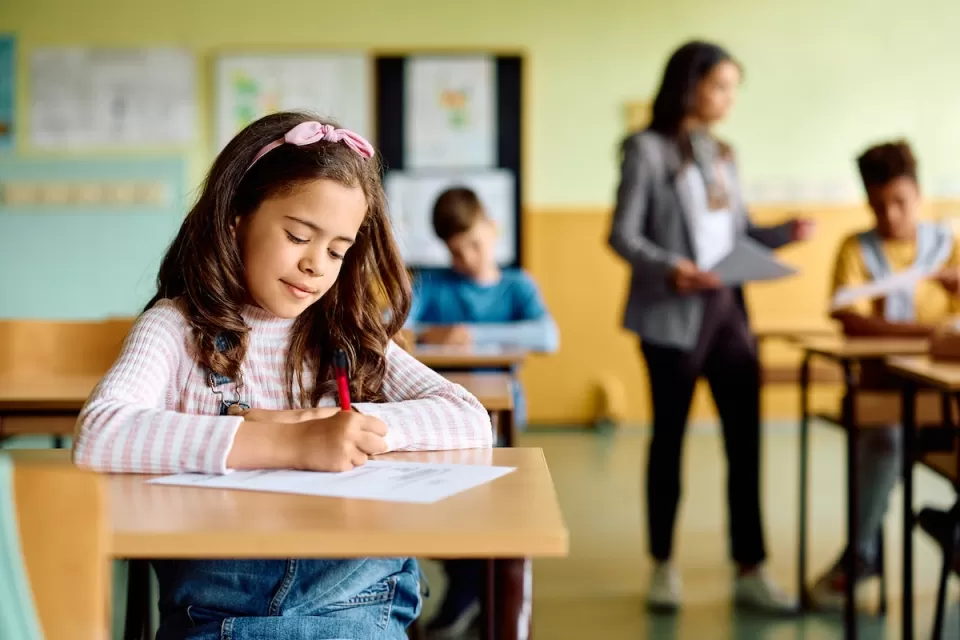 Young girl sitting at school desk taking test with blurred teacher and other students in background.